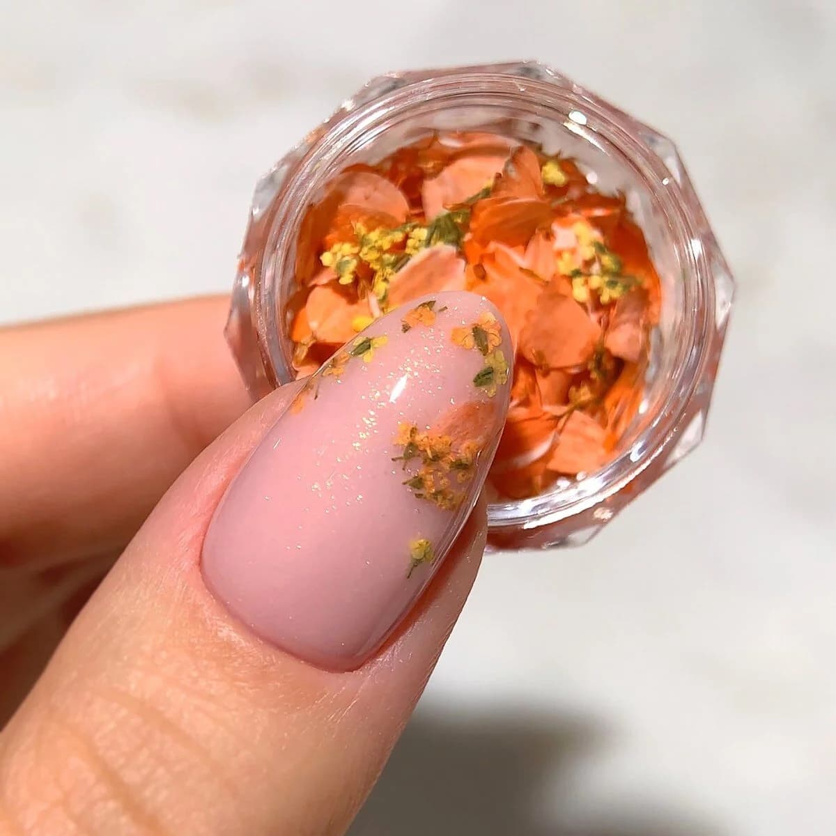 Un pot rempli de pétales de fleurs séchées dans des nuances chaudes dorées et orangées - Semilac Flower Flakes Golden Sunset. Un ongle avec un vsp rose pailleté est décoré de quelques pétales.